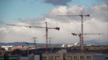 İnşaat vinçleri, binaları ve yapıları, arka planda dağları ve uçan bulutları olan bir şehir manzarası.