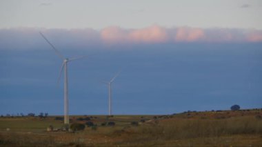 Çerçevenin sol tarafında elektrik jeneratörlü iki rüzgar türbini gün batımında bir tarlada.