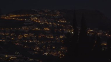 Bu video, tepedeki bir gece şehrinin nefes kesici görüntüsünü yakalıyor. Önünde parlayan küçük evler ve koyu yeşil ağaçlar var.