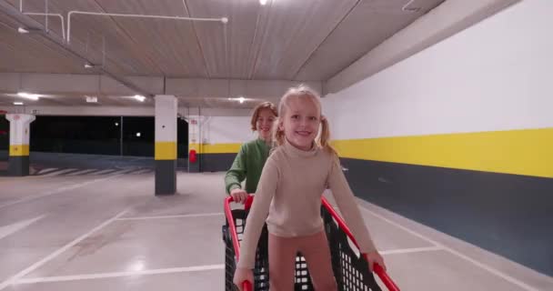 ショッピングカートに座っている5歳の女の子と兄と妹のデュオは 兄としてショッピングセンターの駐車場で爆発しています — ストック動画
