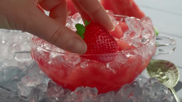 靠近一个透明的玻璃碗 站在冰块上 塞满了西瓜和冰块 女人的手把一个草莓放在一个装有甜点的碗里 — 图库视频影像