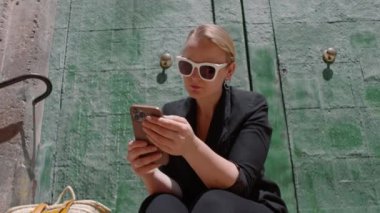 Genç bir kadının beyaz çerçeveli güneş gözlüğü taktığı, büyük yeşil bir kapının yanında elinde cep telefonuyla oturduğu orta boy bir fotoğraf.