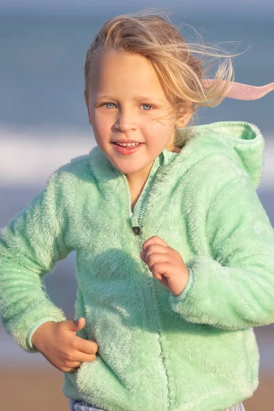 一个穿着绿色夹克 长着蓝眼睛的5岁金发女郎正在跑步 这张长长的照片是她的竖向照片 — 图库照片