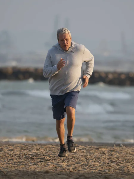 ロングショット 灰色のジャケットとショートパンツの高齢者の垂直写真は 海辺に沿って実行されているアクティブなライフスタイルをリードしています — ストック写真
