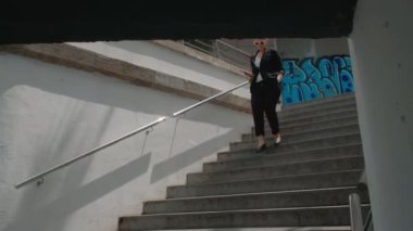 Siyah takım elbiseli genç bir kadın yer altındaki yaya geçidine iniyor ve cep telefonundan bir şeyler okuyor.