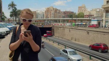 Beyaz çerçeveli güneş gözlüklü genç bir kadın işlek bir caddede elinde cep telefonuyla tünelin yanında duruyor.
