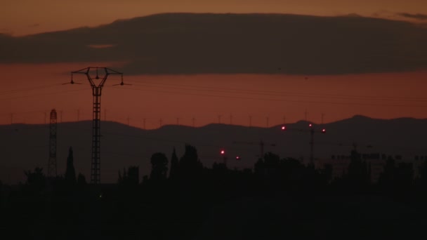 日没時の電力線 遠くの丘の上に立つ木や風力タービンの背景に電柱や高圧線 — ストック動画