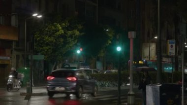 VALENCIA, İSPA - 23 Mayıs 2023: bir kadın otobüs durağında kendini yağmurdan koruyan bir şemsiyeyle bekliyor. Sokak lambaları ıslak yolu aydınlatır. Trafik ışıkları ışıl ışıl parlıyor.