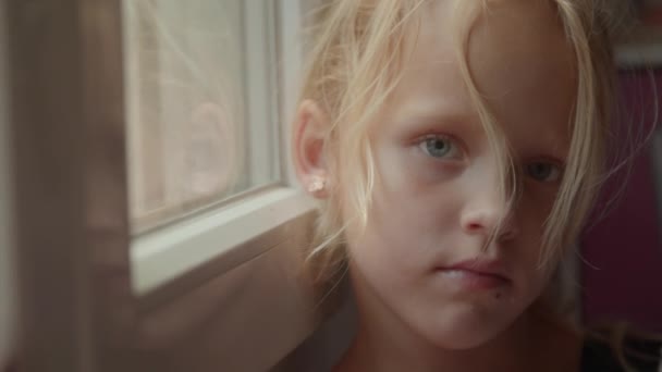 クローズアップショットで 窓のそばに立っている青い目を持つ白人ブロンドの少女の顔 — ストック動画
