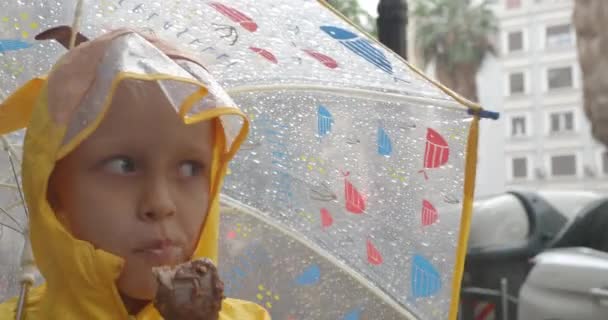 6歳の金髪の少女が黄色いレインコートを着て通りを歩き 豪雨から傘の下に避難し チョコレートアイスクリームを食べる — ストック動画