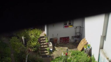 Bahçesi, kırmızı bisikleti, saksısı ve verandaya çıkan merdivenleri olan güneşli bir avlunun penceresinden bak.