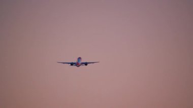 Kırmızı ve beyaz uçak, merkezde, gökyüzünün arka planına karşı pembe bir renkle, irtifa kazanarak tasvir edilir.