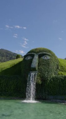 WATTENS, AUSTRIA - 13 Temmuz 2023: Avusturya 'daki Swarovski Müzesi' nde heykel yüzlü ve şelaleli yeşil bir tepenin dikey videosu