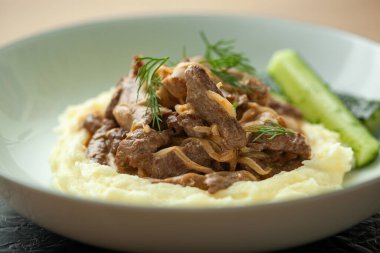 Bir tabak biftek stroganoff yanında patates püresi ve salatalık dilimleri ile servis ediliyor. Yemek taze dereotuyla süslenmiş.