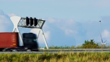 Arka planda açık bir gökyüzü, rüzgar türbini ve yol altyapısı olan bir otoyolda elektronik gişe tahsilat sisteminin altından geçen siyah bir kargo kamyonu görülüyor.