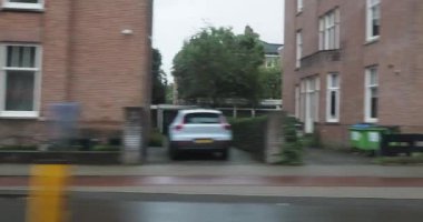 Geleneksel Amsterdam kanal evlerinin önünden geçen arabalar yağmurlu bir günde canlı bir sahne yaratıyor, şehir cazibesini sergiliyor.
