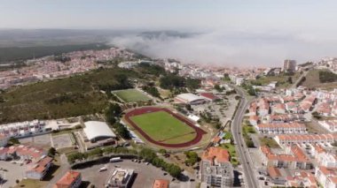 Alçak bulutlar bir stadyumun yer aldığı bir kıyı şehrine doğru hareket ediyor, şehir manzarası üzerinde dramatik bir gökyüzü yaratıyor.