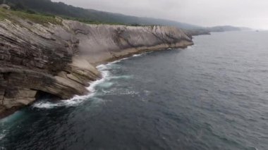 Okyanus dalgaları bulutlu bir gökyüzü altında engebeli kayalıklara çarpar ve sahil boyunca dramatik bir deniz manzarası yaratır.