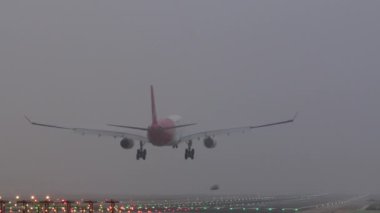 Genişletilmiş iniş takımlı kırmızı ve beyaz uçak sisli koşullarda yanan bir piste yaklaşıyor.