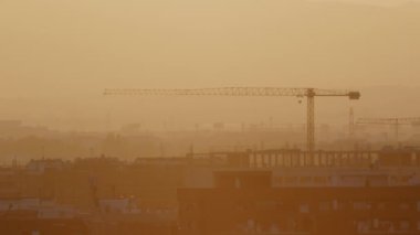 İnşaat vinçleri, şehir gelişimini sembolize eden puslu bir günbatımı arka planına karşı ufuk çizgisine hakim.