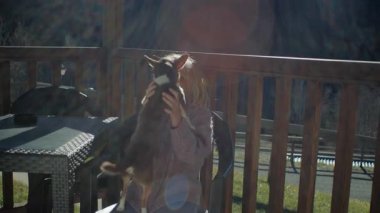 Tahta sandalyede oturan sarışın kız, sıcak güneş ışığı altında huzurlu dağ evi terasının tadını çıkarırken sevgili Chihuahua köpeğine sarılıyor. Eski mercekle çekilen