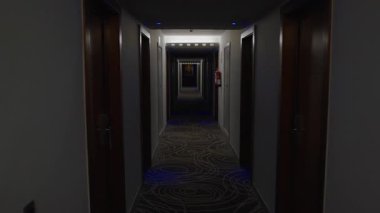 Uzun, karanlık bir otel koridorunun perspektifini yakalamak, kapalı kapılar ve parlayan mavi ışıklar desenli halıyı aydınlatıyor, gizem ve belirsizlik hissi uyandırıyor.