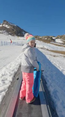 Alp kayak merkezinde kış tatili, renkli kışlık giysiler giyen genç bir kız, karlı dağ manzarasının ortasında yokuş yukarı uçan sihirli bir halı sürüyor.