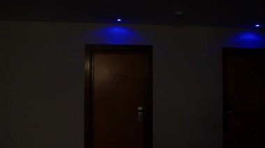 Loş mavi aydınlatma, ıssız otel koridorunda tüyler ürpertici bir şekilde parıldıyor, 220 numaralı odanın yakınında belirsiz bir hoş geldin işareti ile uğursuz gölgeler bırakıyor.