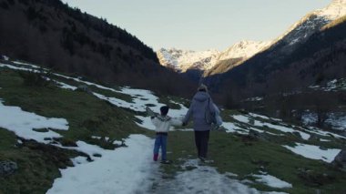 Anne ve çocuk el ele yürüyorlar, Pireneler Ulusal Parkı 'nın karlı dağları arasında, kış manzarasında huzurlu aile bağlarını paylaşıyorlar.