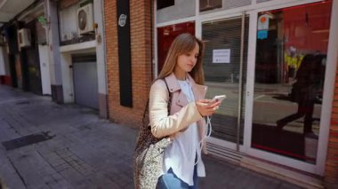 Genç sarışın öğrenci güvenli bir şekilde şehir kaldırımında yürüyor, akıllı telefonuyla meşgul olurken güneşli bir günde kulaklıkla müzik dinliyor.