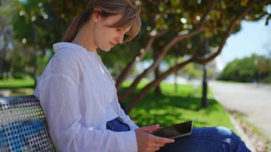 Genç profesyonel kadın güneşli park ortamının keyfini çıkarıyor, tablet aracılığıyla dijital içeriği tarıyor ve dışarıda huzurlu bir an yaşıyor.