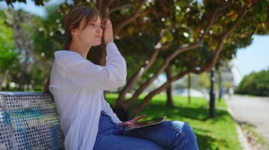 Profesyonel olarak ahşap bankta oturmak, tablet üzerinde uzaktan çalışmak, odaklanmış konsantrasyondan parlak güneş ışığı altında artan hayal kırıklığına kadar değişen yüz ifadeleri sergilemek.