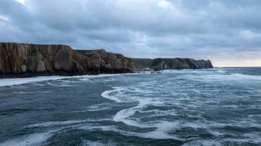 Alacakaranlıkta kıyıya vuran karanlık ve kasvetli okyanus dalgaları Praia da Adraga plajında kayalıklar, Sintra, Portekiz