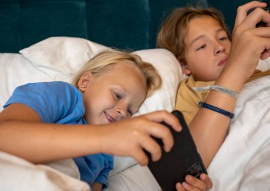 İki kardeş yatakta uzanıyor, akıllı telefonlarıyla online oyun oynuyorlar, rahat bir sabah atmosferinde gülüyorlar ve gülüyorlar.
