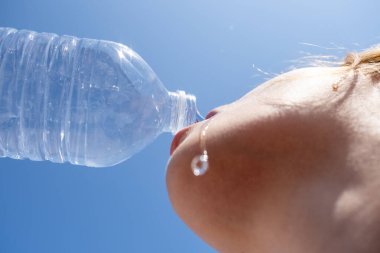 Genç bir kadın sıcak bir günde susuzluğunu gidermek için açık mavi bir gökyüzünün altında plastik bir şişeden ferahlatıcı suyun tadını çıkarıyor.