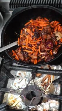 Yemek pişirme süreci taze et ve canlı havuçların gaz sobası üzerindeki dökme demir bir tencerede karıştırıldığını gösteriyor. Dinamik bir mutfak ortamındaki malzemelerin zengin dokularını ve renklerini vurguluyor.