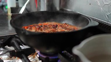 Yemek pişirme süreci, gaz sobasında kaynayan lezzetli bir yemekle dolu dökme demir bir tavayı gösteriyor, canlı malzemeleri vurguluyor ve buhar yükseliyor, davetkar bir mutfak atmosferi yaratıyor.