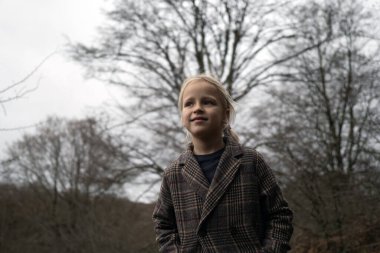 Rahat bir palto giyen genç bir çocuk bulutlu bir kış gününde sakin bir ormanda ağaçların çıplak dallarına bakıyor.