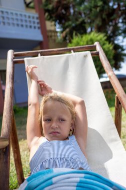 Sarışın kadın iskemlede dinleniyor, ılık güneş ışığında güneşleniyor, yeşillik ve huzurlu yaz atmosferiyle çevrili.