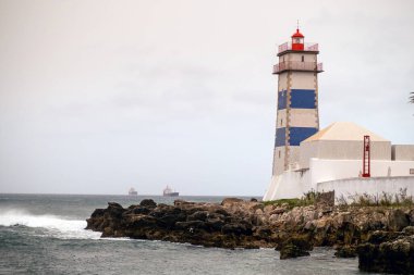 Şeritli deniz feneri kayalık sahil şeridine tünemiş, deniz feneri dalgalar ve sisli deniz manzarası boyunca dalgalanan deniz geçidini aydınlatıyor.