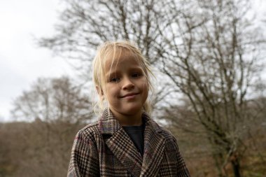 Kareli ceket giyen, kışın ormanda ağaçların önünde poz veren gülümseyen sarışın bir çocuğun portresi.