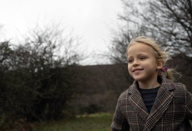 Sarışın kız yapraklı parkta yürüyor, bulutlu bir sonbahar öğleden sonrasında kareli ceket giyiyor, sakin bir an yaşıyor.