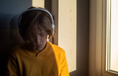 Kulaklık takan bir genç, bir pencerenin yanında otururken müziğe dalmış, rahatlatıcı doğal ışığın tadını çıkarıyor ve yalnızlığın tadını çıkarıyor.