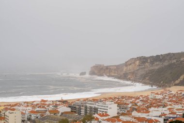 Nazare sahilinde dalgalar kıyıya vuruyor. Arka planda, bulutlu bir gökyüzünün altında dramatik uçurumlar yükseliyor.