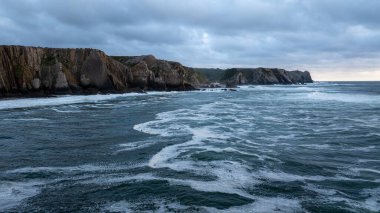Karanlık okyanus dalgaları alacakaranlıkta kayalık bir kıyı şeridine çarpıyor, Sintra, Portekiz 'de dramatik ve karamsar bir deniz manzarası yaratıyor.