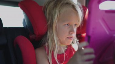 Kırmızı araba koltuğunda oturan sarışın kız odaklanmış ifadesiyle pembe tableti izliyor.