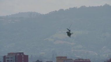 Tek bir helikopter arka planda sisli dağ manzaralı binalar üzerinde keskin bir sahile çıkıyor.