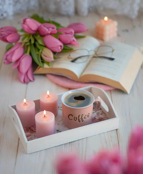A cup of coffee with a book and glasses, tulips, candles in the home interior, an aesthetic photo