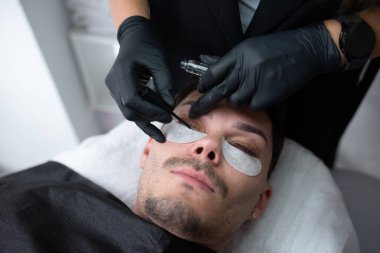 Erkek müşteri, görünümü geliştirmek için hassas araçlar kullanan yetenekli bir teknisyenle çağdaş bir güzellik salonunda kirpik uzatma uygulamasından geçiyor.