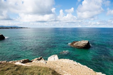 Kıyı manzarası kayalık kıyıları, canlı turkuaz suları ve yukarısında tüylü bulutları sergiliyor. Rahatlamak ve doğayı takdir etmek için mükemmel bir atmosfer yaratıyor.